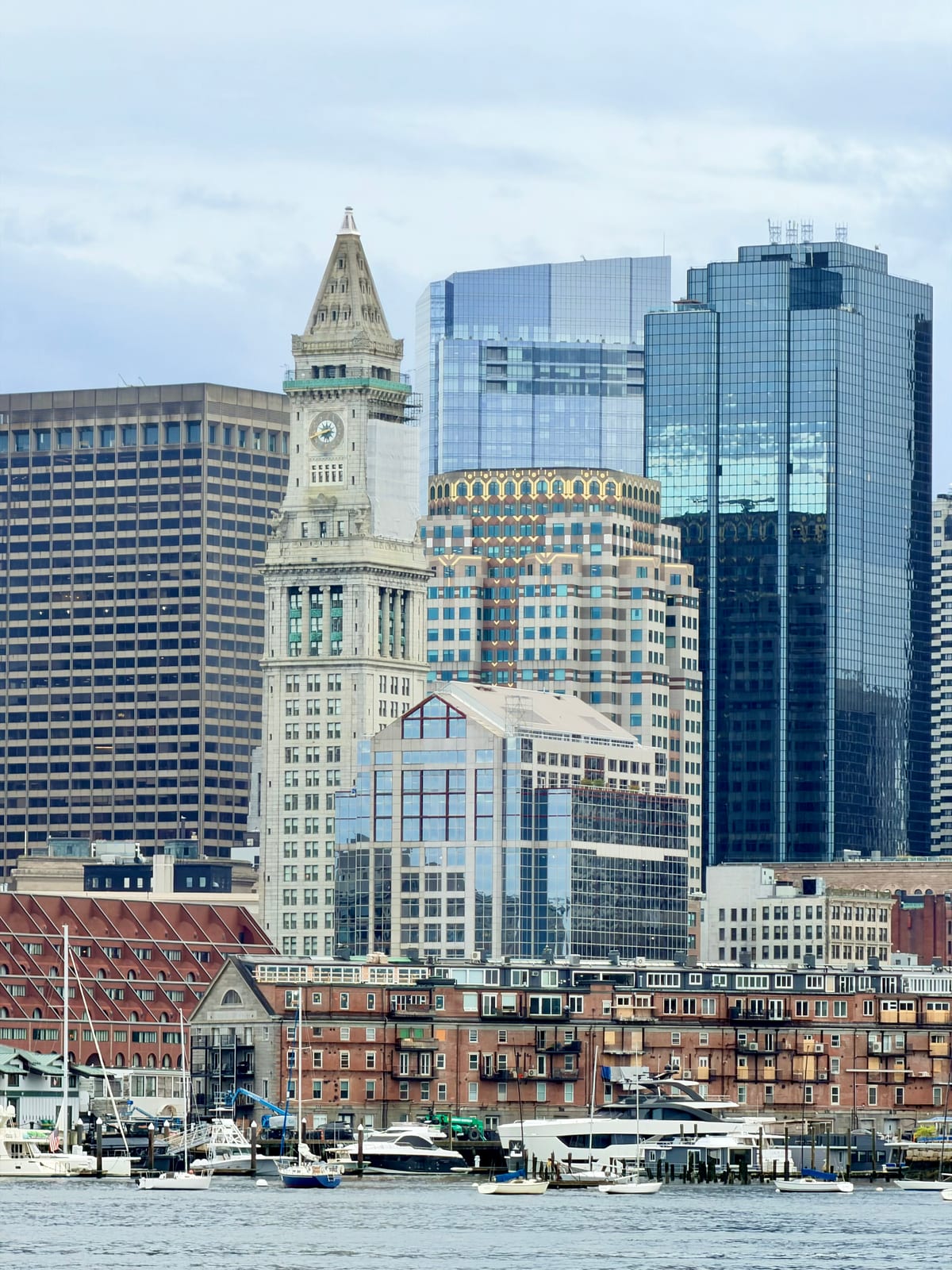 A stone clock tower and glass skyscrapers above brick waterfront buildings and boats in Boston Harbor