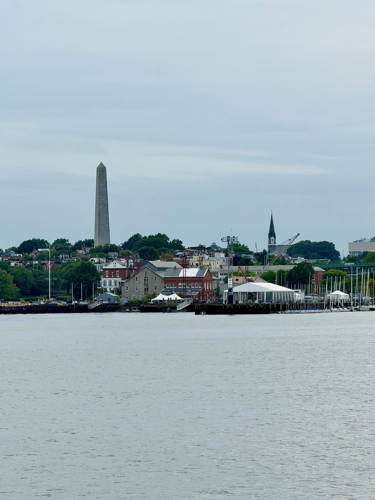 A stone obelisk and church steeple rising above waterfront buildings and sailboat masts across the water