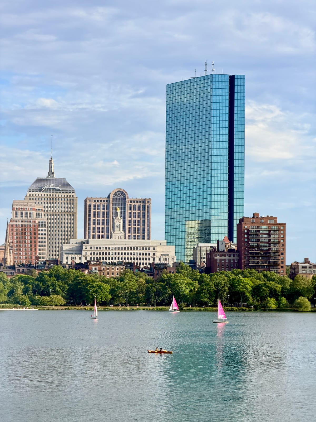 Pink-sailed boats and a yellow kayak on the Charles River below Boston's Back Bay skyline and a tall blue glass tower