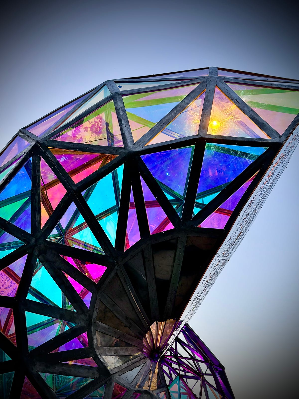 An angular outdoor sculpture with iridescent triangular panels glowing pink, blue, green, and gold in a dark metal frame